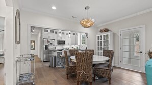 A modern kitchen with white cabinets, a central island, and a dining area with wicker chairs. - Cottage Glyn - 3 BR Home Steps to the Beach (North Myrtle Beach)