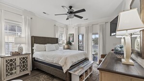 A bedroom with a large bed, a ceiling fan, a bedside table with a lamp, a dresser, and a view of the outdoors. - Cottage Glyn - 3 BR Home Steps to the Beach (North Myrtle Beach)