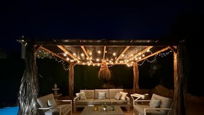 Een buitenzitgedeelte met een houten pergola, lichtslingers en een zwembad.