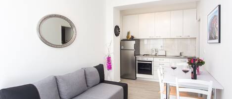 Una sala moderna con un sofá gris, una mesa de comedor con sillas blancas, una zona de cocina con armarios blancos y un espejo redondo en la pared.