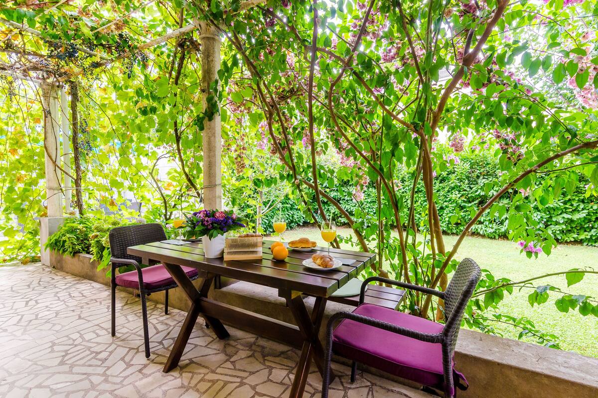 An outdoor patio with a table set for two, surrounded by lush greenery and a pergola.