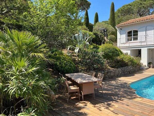 Un espace aménagé au bord de la piscine, avec une terrasse en bois, une table à manger entourée de chaises et un bâtiment blanc doté d’un balcon.