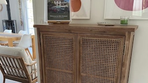 Un meuble en bois avec un panneau avant tissé, une pancarte d’accueil encadrée, et un coin repas avec des chaises et une table en arrière-plan.