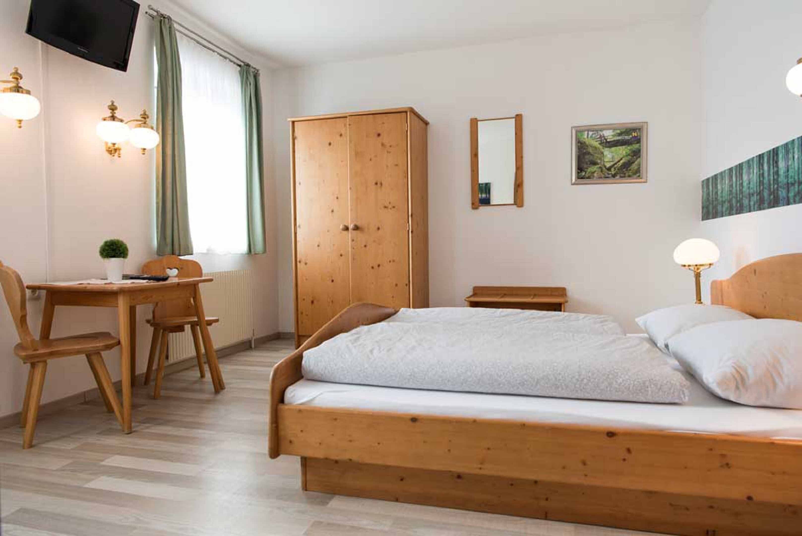 A bedroom with a wooden bed, a wooden wardrobe, a desk with a chair, and a television mounted on the wall.