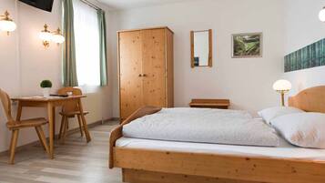 A bedroom with a wooden bed, a wooden wardrobe, a desk with a chair, and a television mounted on the wall.