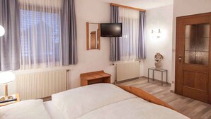 A hotel room with a bed, a TV mounted on the wall, a wooden nightstand, a small table, and a door with a frosted glass panel.
