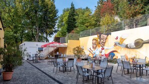 An outdoor café with tables and chairs, a mural on the wall, and a staircase leading to an upper level.