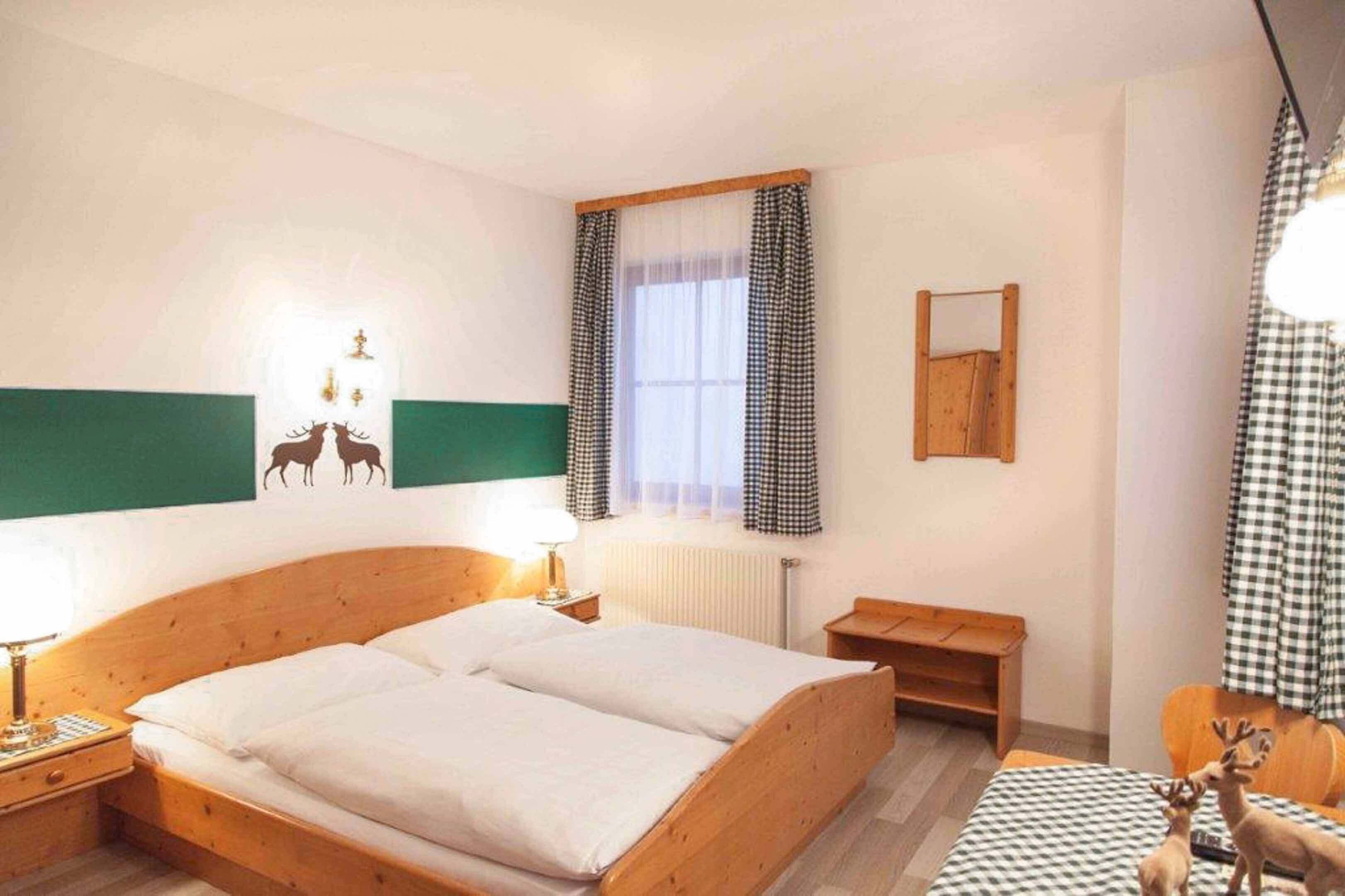 A bedroom with a wooden bed, a nightstand, a small table, a window with checkered curtains, and wall decorations including a lamp and a mirror.