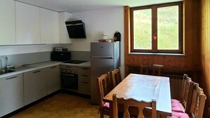 A compact kitchen with a dining table and chairs, a refrigerator, and a window with a view of greenery. - Sweet Home ski out (Valtournenche)