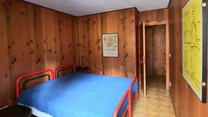 A small, cozy room with wooden walls and a tiled floor. It features a single bed with a blue mattress and red metal frame, a small table with a chair, and a map on the wall. - Sweet Home ski out (Valtournenche)