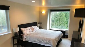 A bedroom with a bed, bedside tables, a lamp, a TV, and a window with a view of trees. - Moden West Seattle House in the Trees (Seattle)