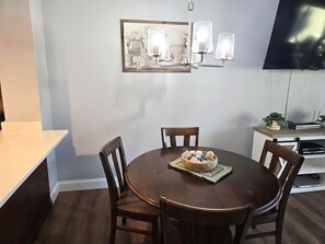 Un comedor con una mesa redonda, cuatro sillas, un centro de mesa, una lámpara de pared y un televisor de pantalla plana sobre un mueble.