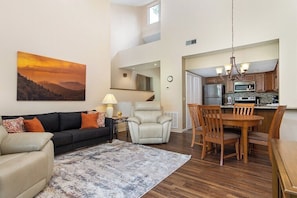 A living room with a sofa, a chair, a rug, a dining table with chairs, a kitchen area with appliances, and a painting on the wall. - Fairway Villa 1501 at Rumbling Bald (Lake Lure)