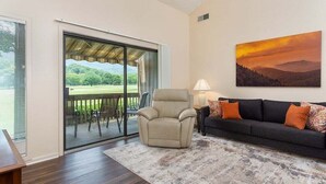 A room with a sliding glass door leading to a deck, a beige armchair, a dark sofa with orange cushions, and a painting of a sunset. - Fairway Villa 1501 at Rumbling Bald (Lake Lure)