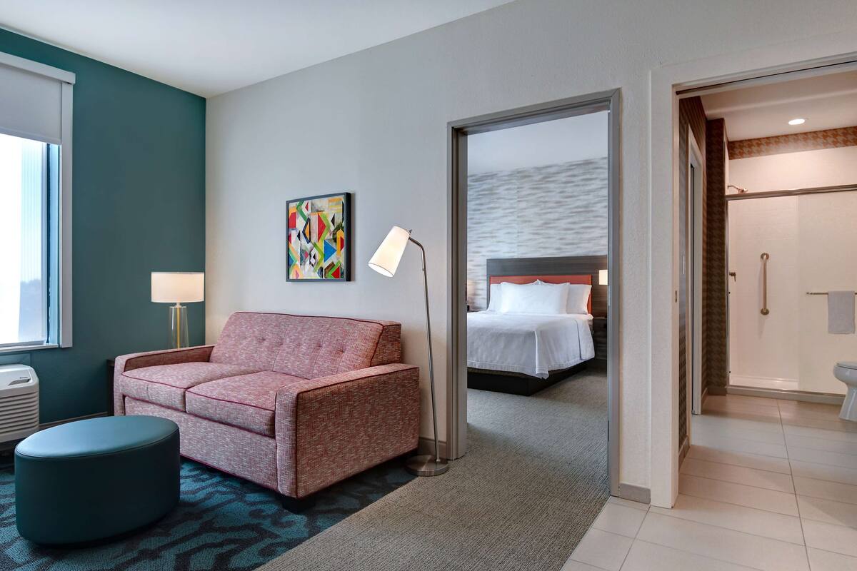 Habitación de hotel con sofá, lámpara, un cuadro, cama y baño visible a través de una puerta abierta.