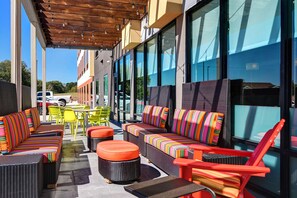 An outdoor seating area with wicker furniture and colorful cushions, large windows, and a wooden pergola. - Home2 Suites by Hilton North Plano Hwy 75 (Plano)