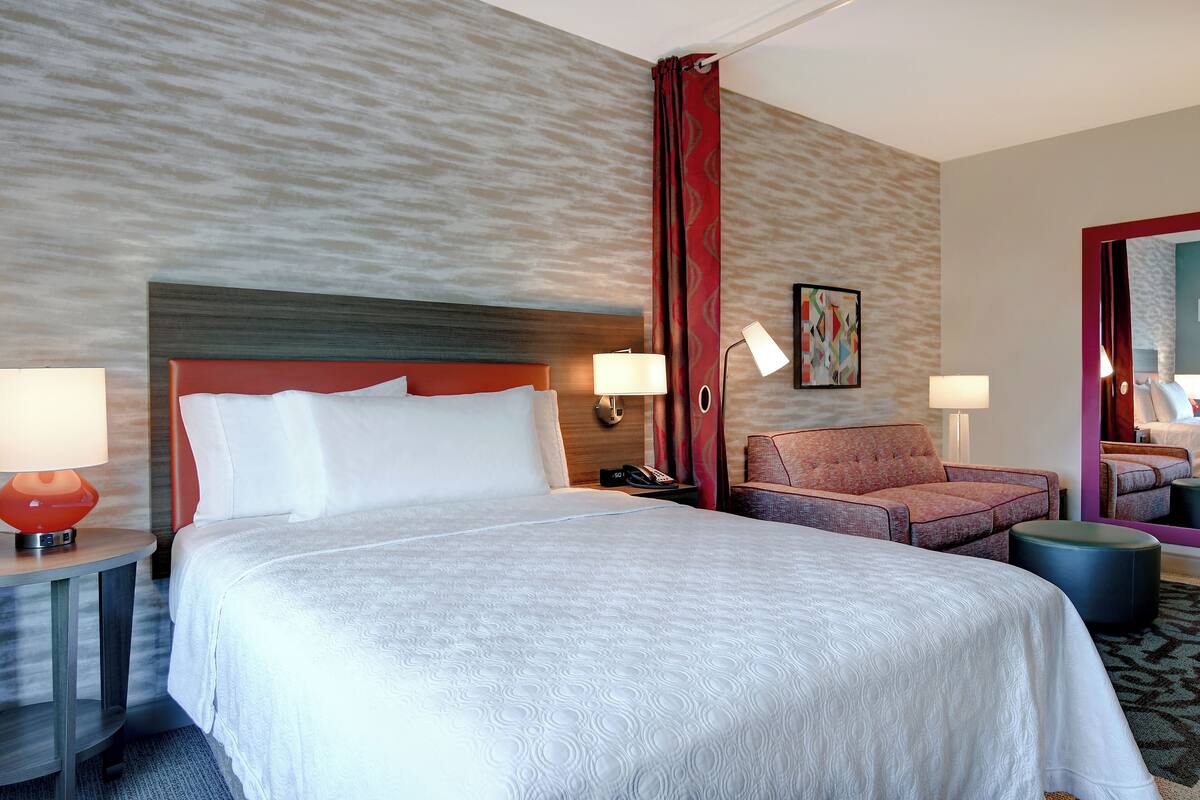 Habitación de hotel con una cama grande, dos mesitas de noche con lámparas, una silla, un pequeño reposapiés y un espejo.