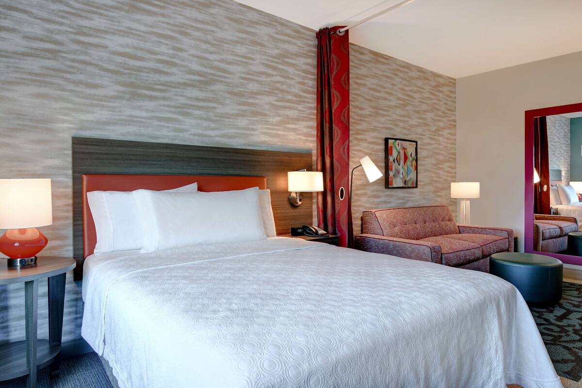 Habitación de hotel con una cama grande, dos mesitas de noche con lámparas, una silla y un pequeño otoman.