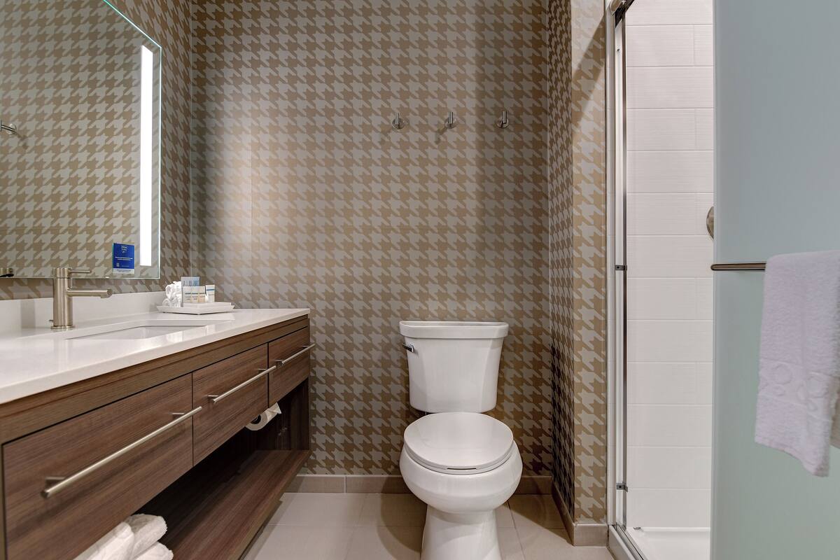 Un baño con pared estampada de pata de gallo, un inodoro blanco, un tocador de madera con espejo y una cabina de ducha de vidrio.