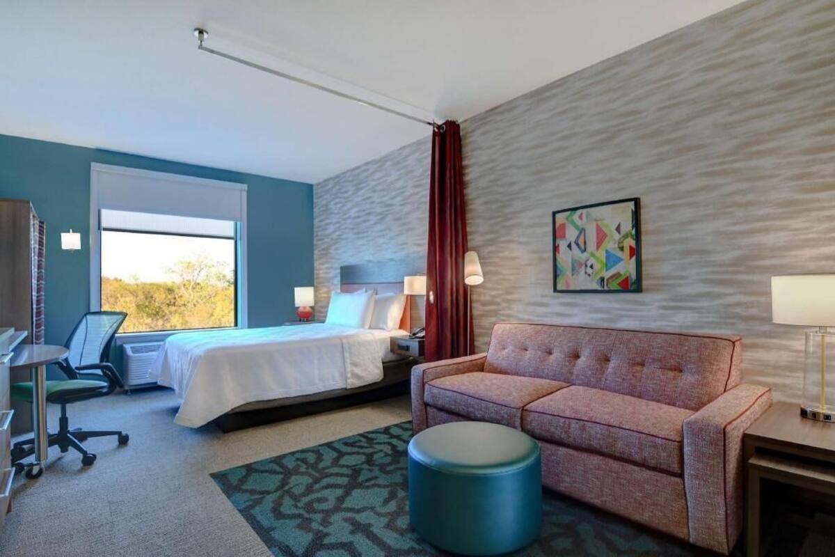 Habitación de hotel con cama, escritorio, silla, sofá, reposapiés redondo y ventana con vistas.
