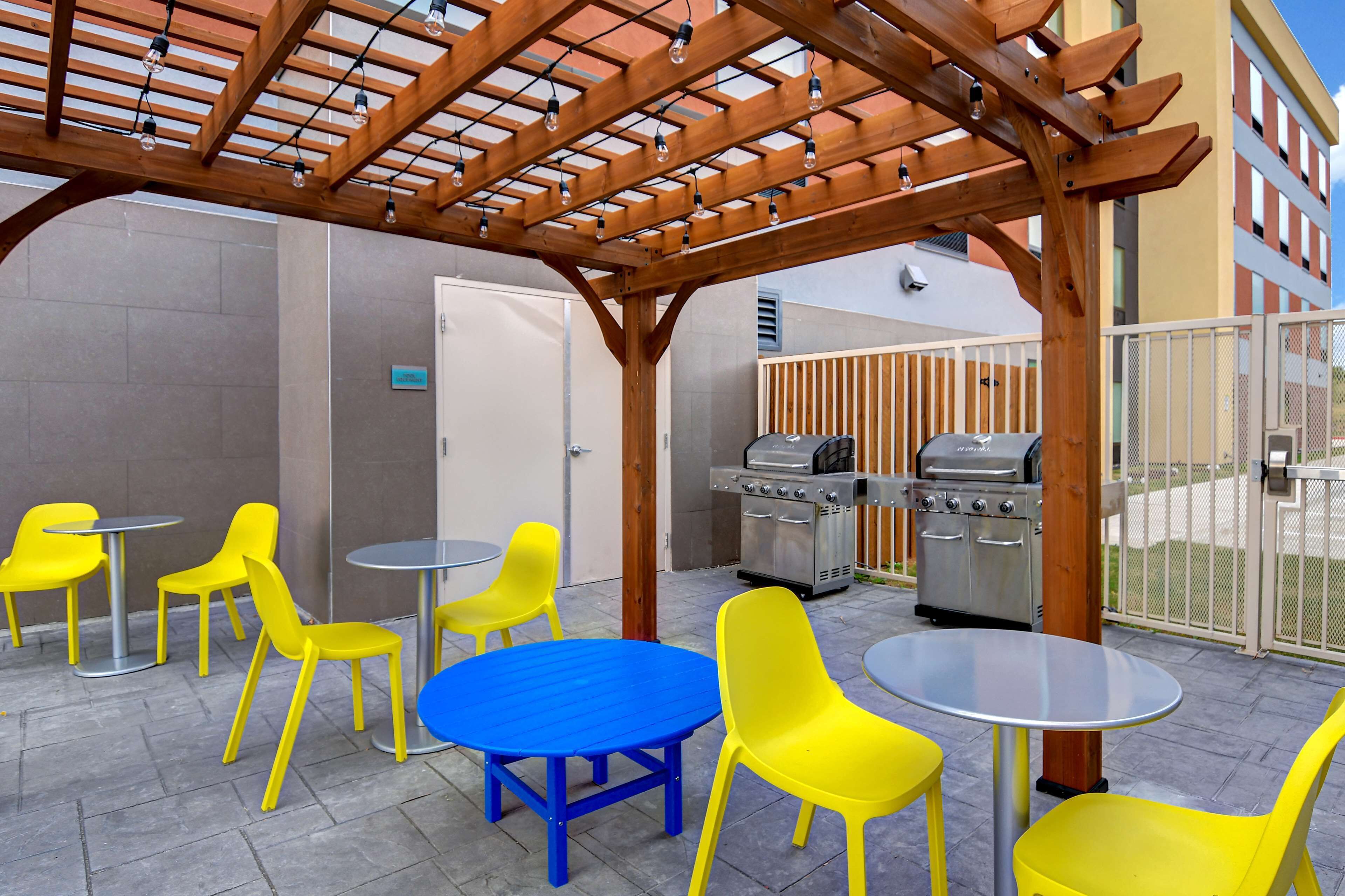 An outdoor patio with yellow chairs and blue tables, a wooden pergola with hanging lights, and a barbecue grill.