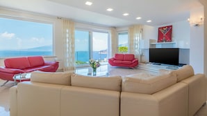 A modern living room with a beige sofa, red armchairs, and a flat-screen TV. Large windows offer a view of the sea and a distant mountain. - CASA ANGELICA LUXURY VILLA (Priora)