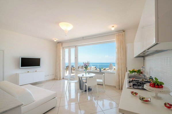 Una sala de estar moderna y bien iluminada, con un gran ventanal, un televisor de pantalla plana, un sofá blanco, un área de comedor con mesa y sillas, y una cocina con estufa y encimera.