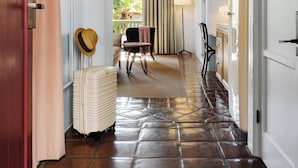 A hallway with a tiled floor, a wicker ceiling light, a door on the left, and a chair with a table in the background. - Palihouse Santa Barbara (Santa Barbara)