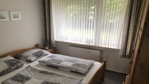Un dormitorio con una cama de madera, dos mesitas de noche, una lámpara, un radiador y una ventana con cortinas translúcidas.