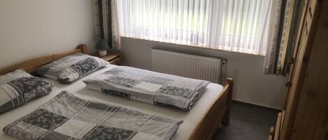 A bedroom with a wooden bed, two bedside tables, a lamp, a radiator, and a window with sheer curtains.