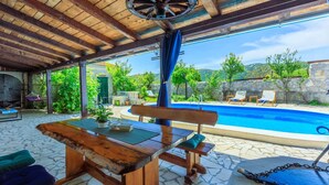 Un área de piscina al aire libre con una mesa y sillas de madera, una hamaca y vistas a la piscina y la vegetación circundante.