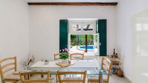 Un comedor con mesa de vidrio, sillas de madera y vistas a una piscina y jardín a través de ventanas abiertas.