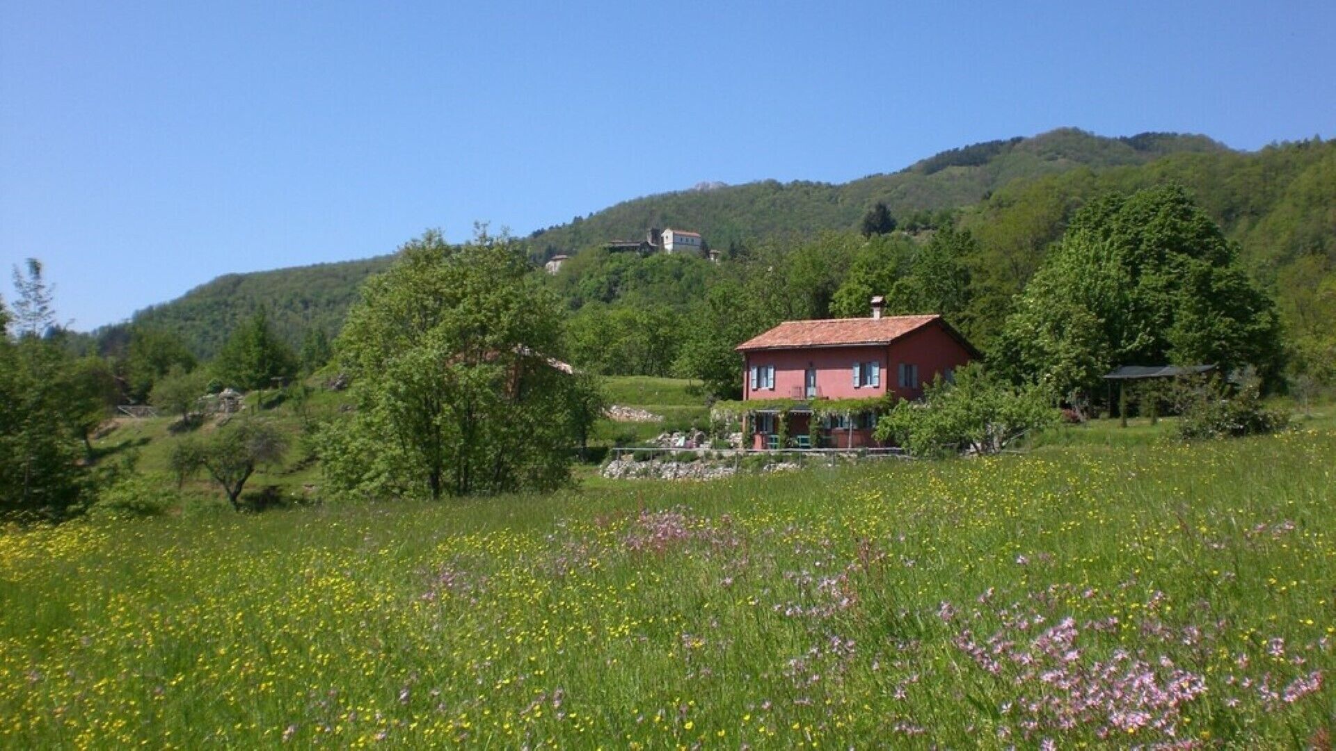 Ein rotes Haus mit Ziegeldach, umgeben von einem Feld mit Wildblumen und Bäumen, vor einer Hügelkulisse.