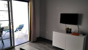 En moderne stue med en skydedør til en balkon, et fladskærms-tv monteret på væggen og et hvidt skab med en plante og en fjernbetjening ovenpå.
