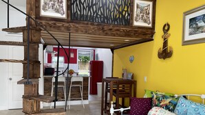 Ein gemütlicher Wohnraum mit einer Holztreppe, einem Sofa mit gemusterten Kissen und einer durchgehenden Tür erkennbaren Küchenzeile.
