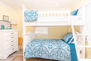 A bunk bed with a patterned bedspread, a dresser with drawers, and a wall-mounted lamp.