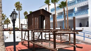 A wooden climbing structure in an outdoor area with a white building in the background.