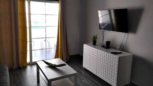 A modern living room with a white sofa, a coffee table, a white cabinet with a textured surface, a flat-screen TV mounted on the wall, and a window with curtains.