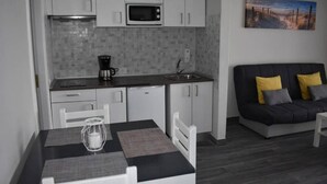 A compact kitchen with white cabinets, a microwave, and a coffee maker. There's a dining table with a placemat and a candle. A black sofa with yellow cushions is against the wall.