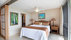 Une chambre à coucher avec un lit, deux tables de chevet, un ventilateur de plafond, un tableau et une fenêtre avec des rideaux.