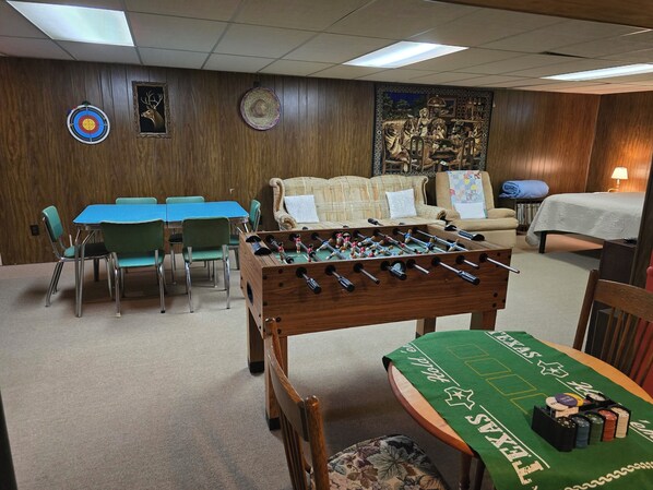 A room with a foosball table, a dartboard, and a painting of animals. There are chairs and a table with a green tablecloth.