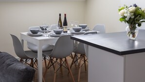 Un coin repas avec une table blanche dressée pour quatre personnes, comprenant des verres à vin, des bols et une bouteille. On y trouve des chaises grises aux pieds en bois, et un vase avec des fleurs sur la table.