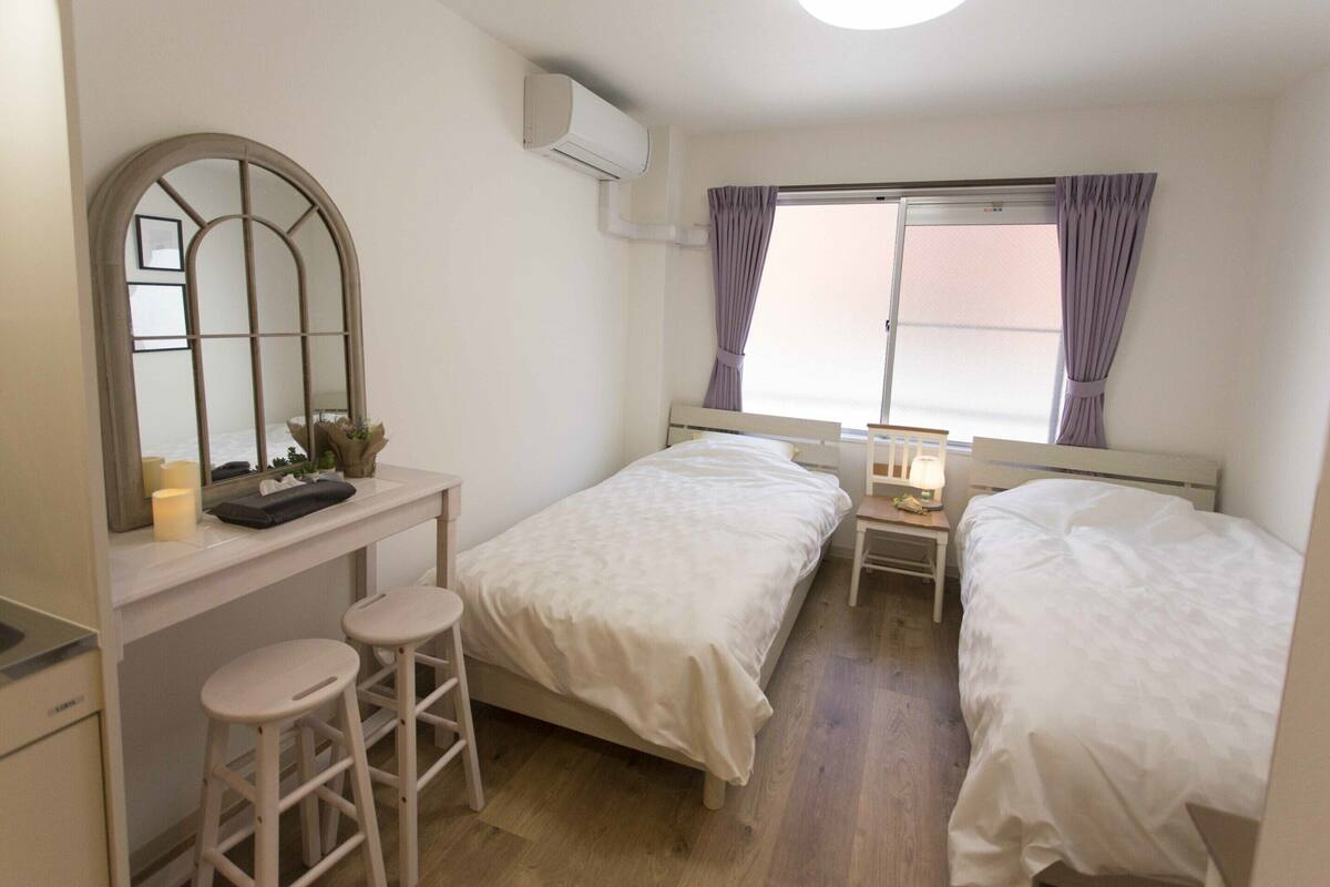 A bedroom with two beds, a mirror, a small table with a lamp, and a window with curtains.