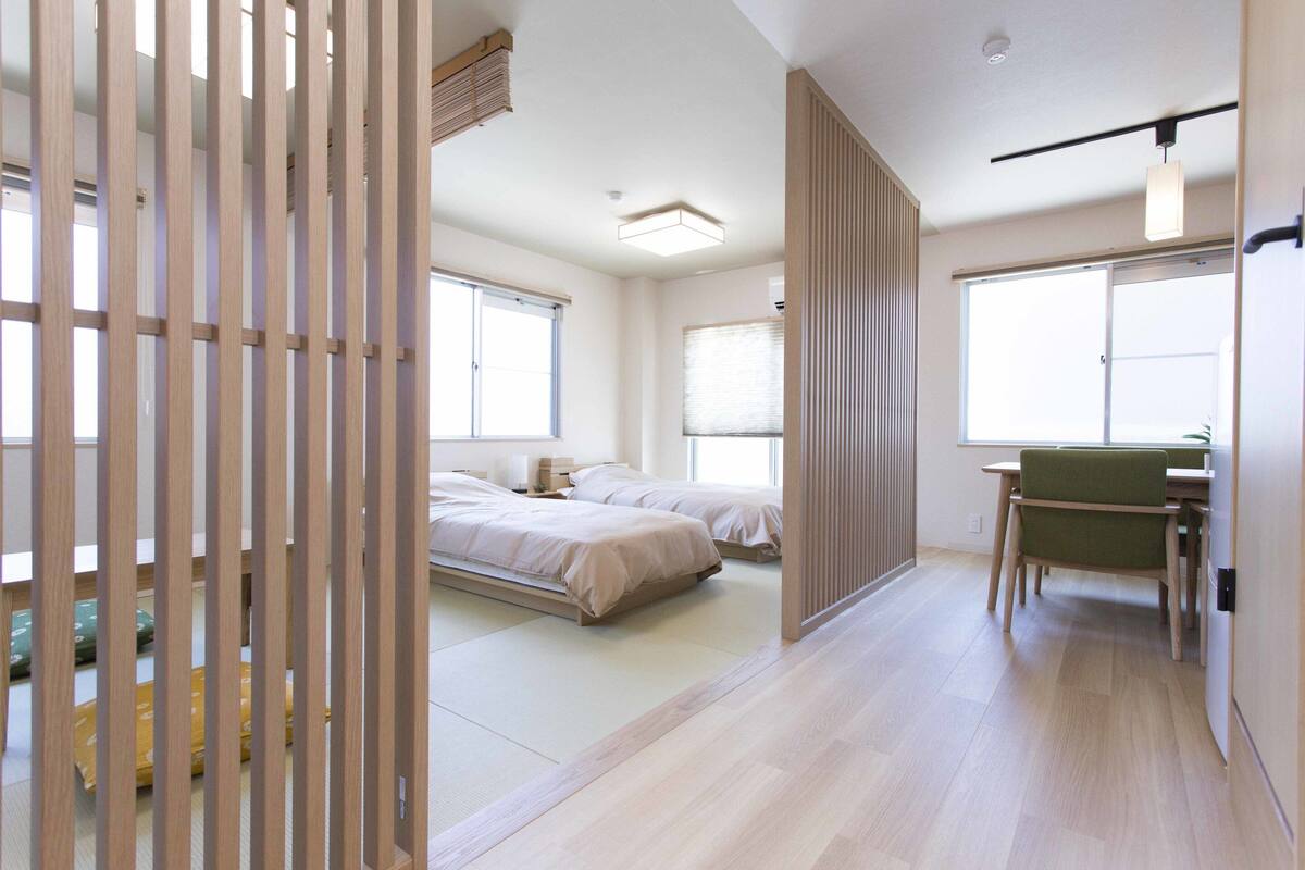 A modern hotel room with a large bed, a desk, and a chair. There are large windows providing natural light.