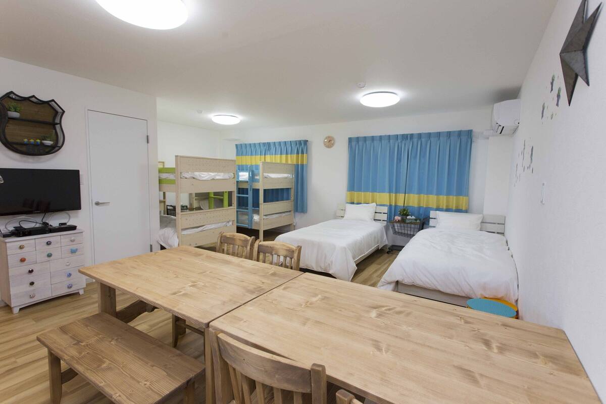 A room with a wooden table, two beds, a dresser, and a television.