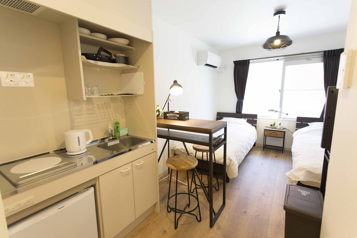 A compact hotel room with a kitchenette, a single bed, a small table with a lamp, and a window with curtains.