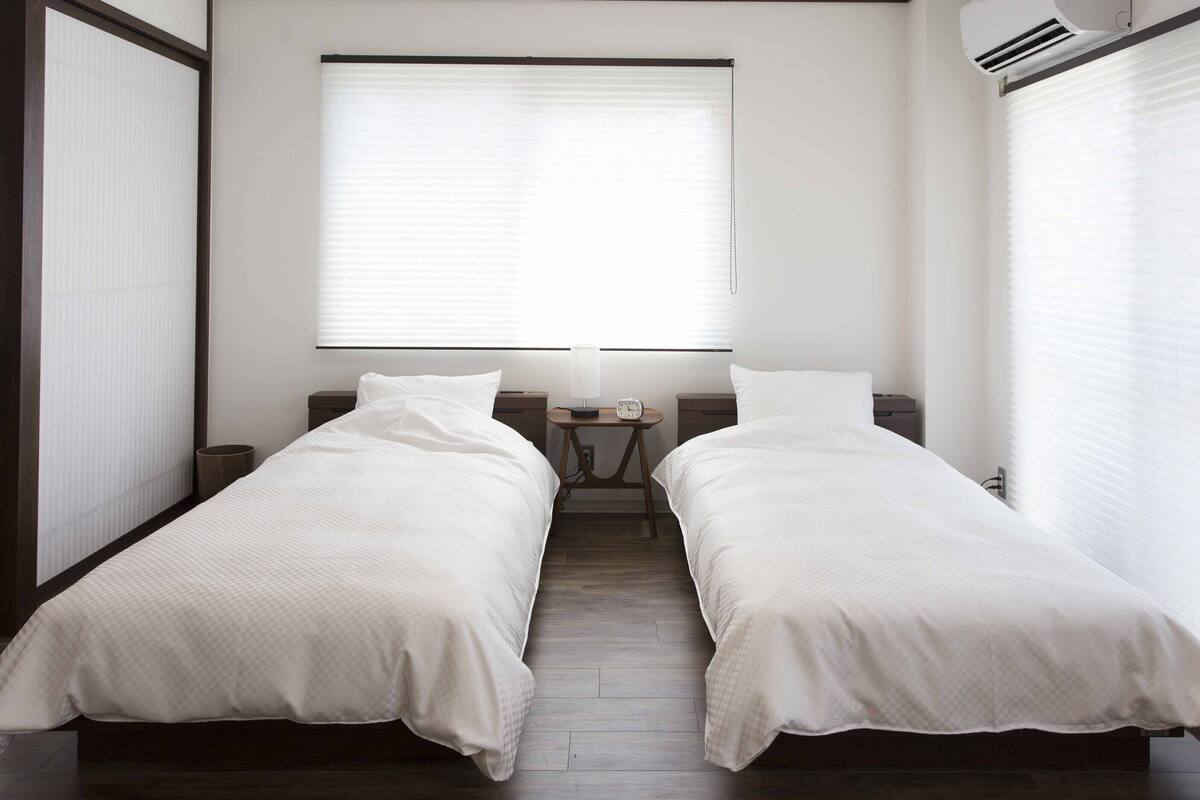 A twin room with two beds, a bedside table with a lamp, and a window with blinds.