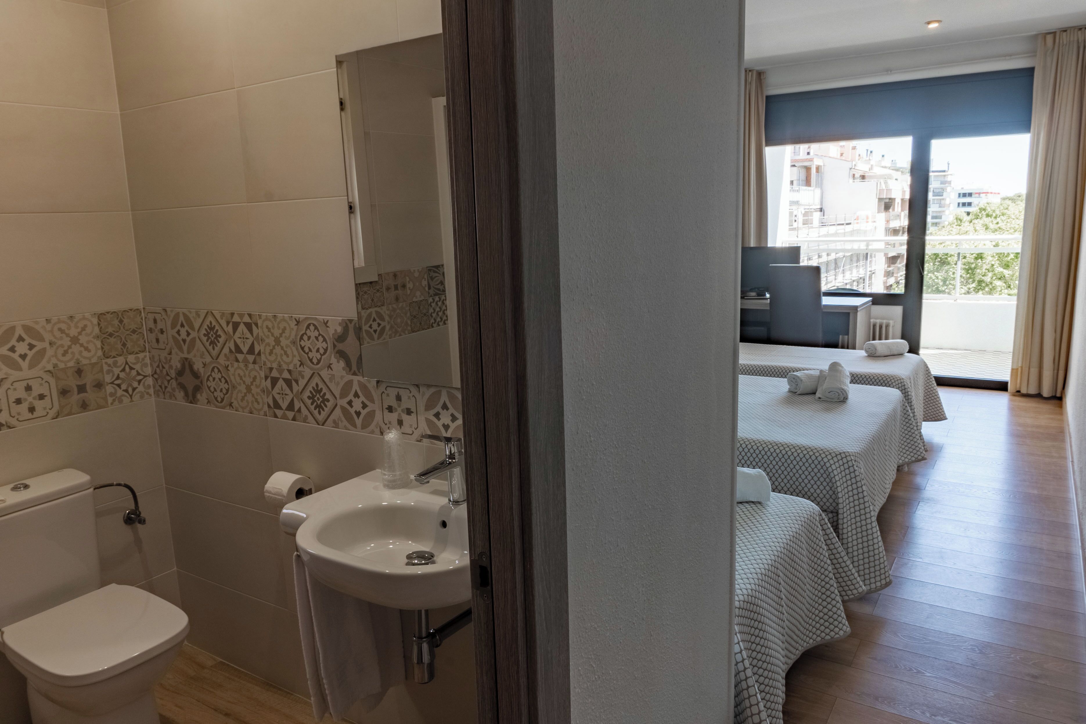 Une salle de bain d’hôtel avec des toilettes, un lavabo et des carreaux décorés. Il y a un balcon avec une table et des chaises.