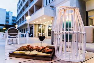 A wooden table with a tray of food and a glass of red wine, a white candle in a glass holder, and outdoor seating with white chairs.