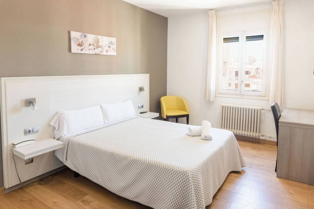 Habitación de hotel con una cama grande, una mesita de noche, una silla, un radiador y una ventana con cortinas.